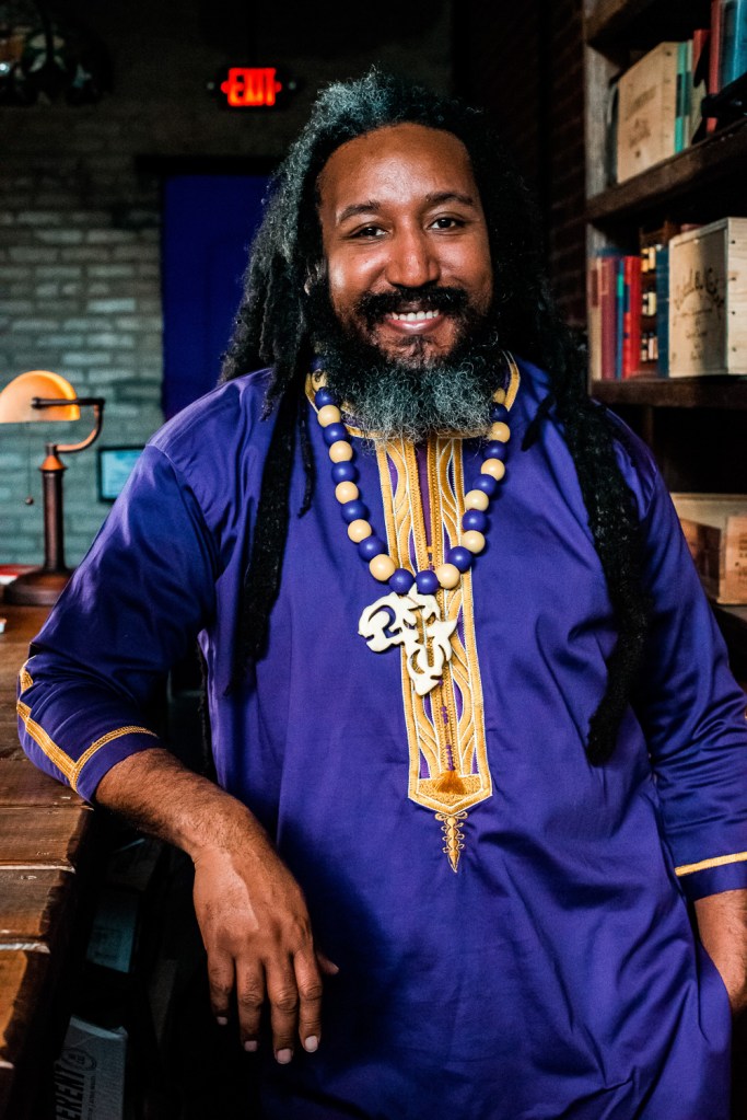 Portrait of Dr. Malik Boykin. Malik stands as a smiling black man with dreadlocks, wearing purple regalia and an African necklace in a library.